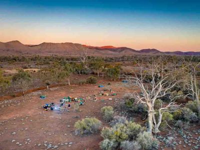 A landscape scene taken at sunset in the Flinders Ranges with soft hues of blues, yellows, orange and reds. There's a group of travellers camped in a clearing with swags and the smoke of a campfire. The ranges are in the background and the camp is surrounded by various bushes and shrubs.