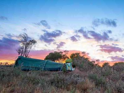 Outback Australia at dawn with a swag bedroll set up in the foreground. Bushes and grasses are scattered around and the glow of sunrise illuminates the outback sky.