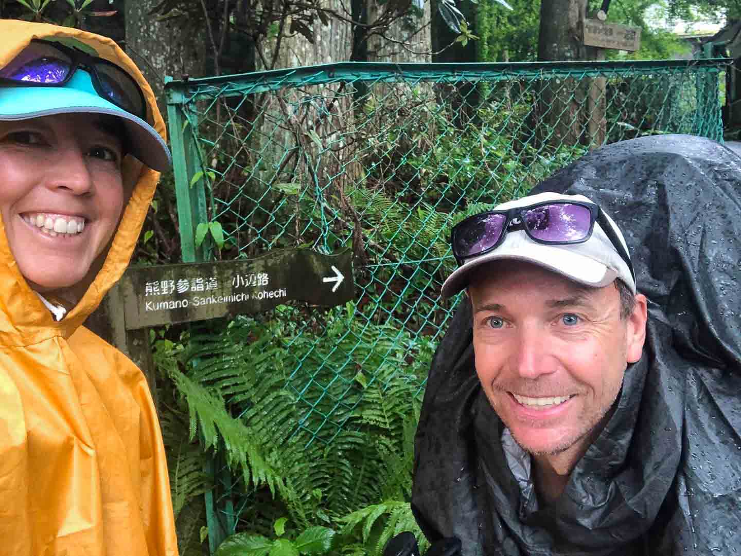A woman in a yellow rain cover takes a selfie with a man wearing a black rain cover. They are both ginning at the camera at the start of the Kohechi Kumano Pilgrimage Route in Japan. They both have matching sunglasses resting on top of their caps and are about to set off for the trek.