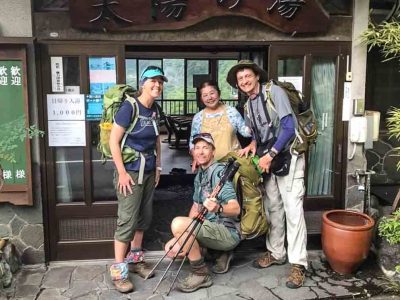 A group of four people, three are travellers and the other is a local Japanese host. They are gathered together smiling at the camera in front of the homestay entrance. The travellers are hikers all wearing backpacks and it looks to be a farewell photo.