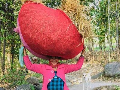 Women's India Wellbeing Journey. An Indian woman dressed in blue pants with a checked blue and dark indigo overdress and bright pink cardigan. She carries a massive bundle of hay on top of her head. The bundle is wrapped in a bright red cloth and she is smiling broadly at the camera.