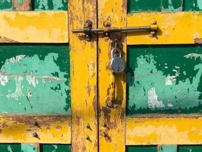A close-up of a rustic door painted in chipped green and yellow paint. There is a bloated latch with silver padlock to secure the opening.