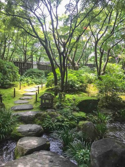 A stepped stoned path leads over a gentle garden stream and off into the distance. It is surrounded by the beautiful green manicured gardens of Koko-en in Japan.