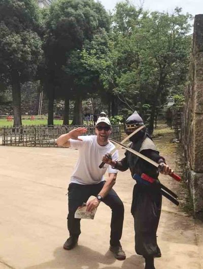 An Aussie tourist dressed in a white t-shirt and dark pants strikes a battle pose next to a Japanese ninja in traditional dress and holding swords in each hand.