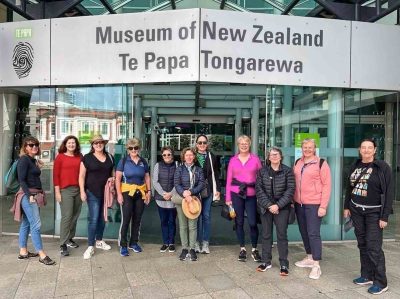 A group of 9 women stand in front of the Te Papa Tongarewa Museum in Wellington. They are all smiling at the camera.