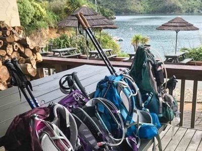 4 large backpacks each with a set of hiking poles rest against an outdoor table on a deck overlooking a remote bay surrounded by lush rolling green hills.