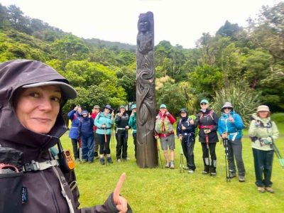 A walking guide is close up in the lower left corner, taking a 'selfie' with her group of 10 women behind her gathered around a tall carved Maori totem.