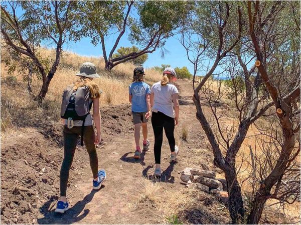 Three tween kids have their backs to the camera and are walking along a bush track near the coast. A few Australian native trees are dotted about but mostly the scene is filled with the dirt track and dry grasses. The teenage girl at the back of the group is wearing a backpack.