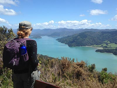 A woman wearing a small backpack and cap stares out across the beautiful ocean inlets and rolling green hills of New Zealand's Marlborough Sounds.